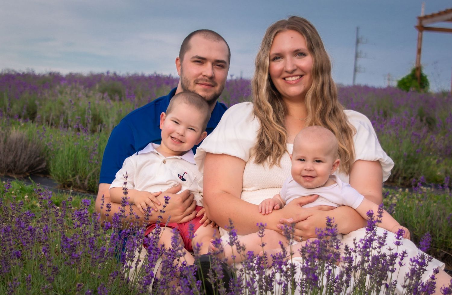 Family of four posing in a blooming lavender field, parents holding two young children, smiling outdoors.