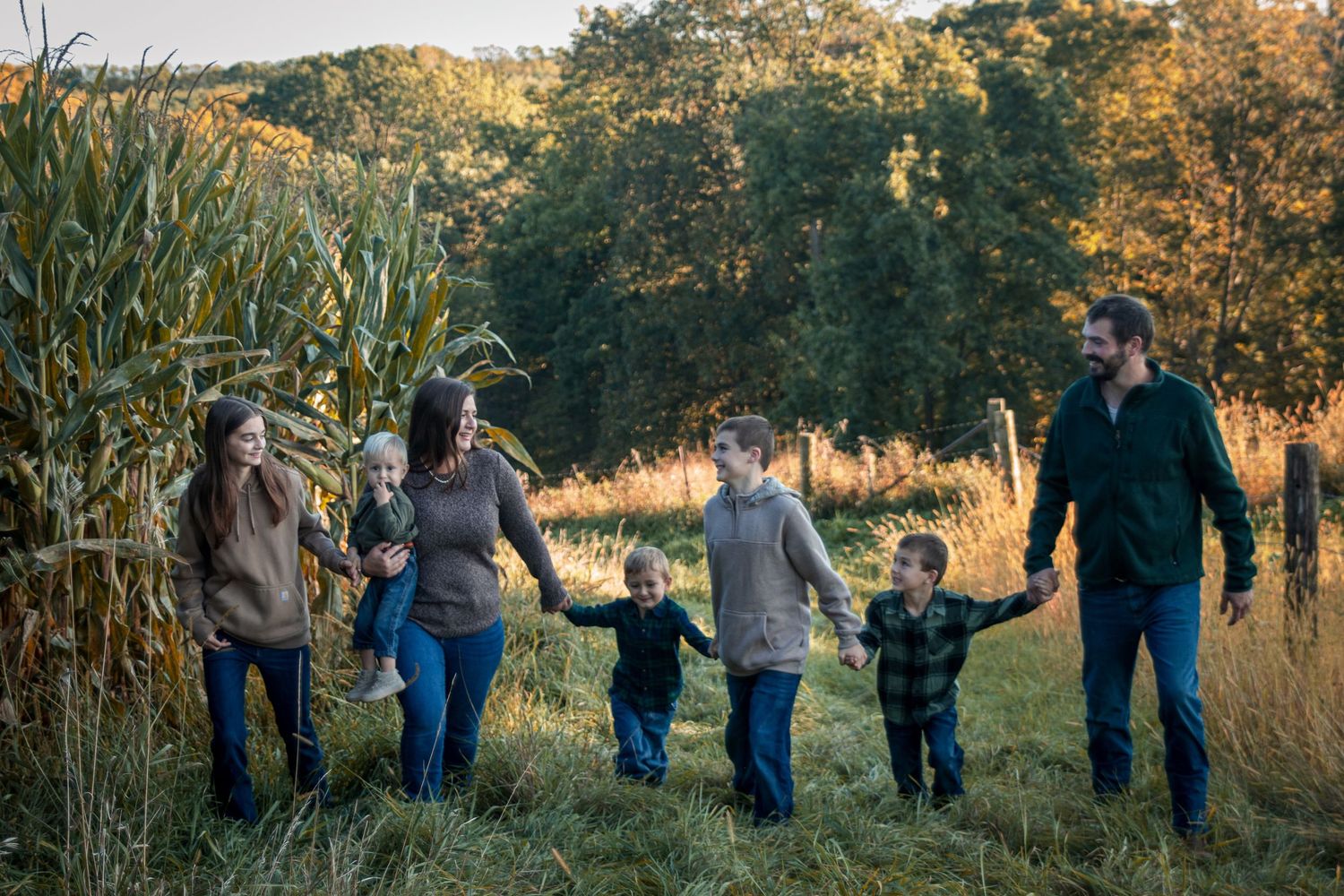 Family of six walking hand in hand through a golden autumn field beside a cornfield at sunset.