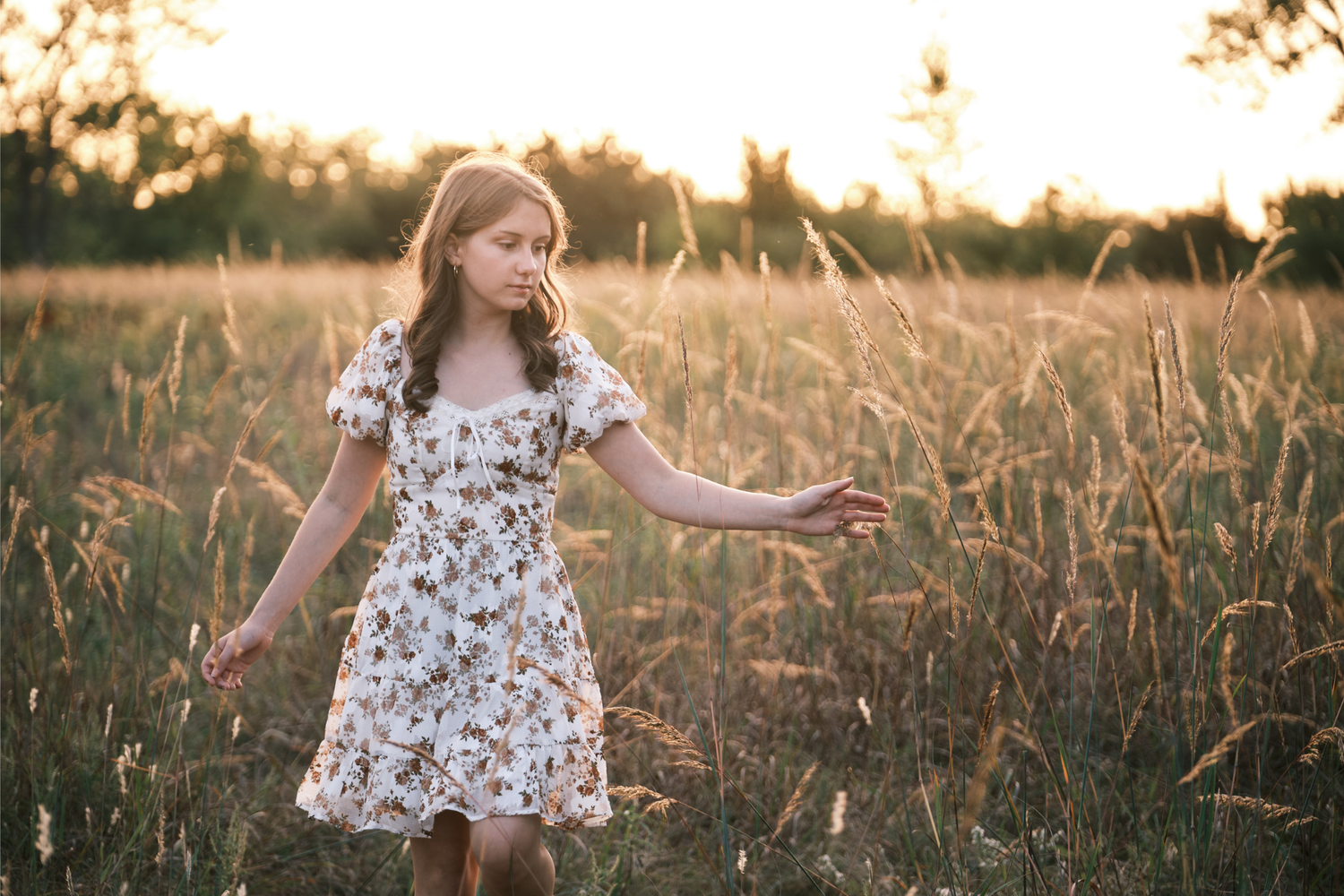 A young girl in a floral dress walks through a golden sunlit field, gently touching tall grass at sunset.