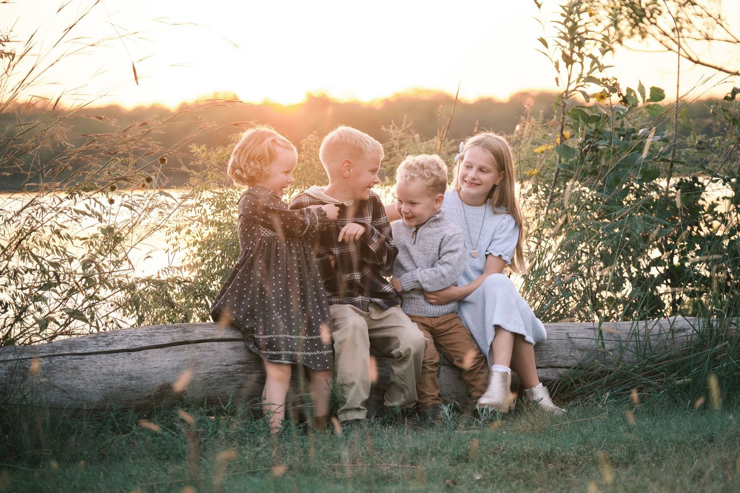 Four happy siblings sitting on a log near a lake surrounded by warm golden sunset light.