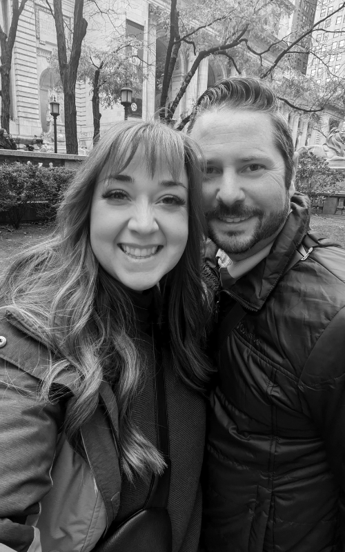 Black and white selfie of owners, Beth + Brian, outdoors in a park with trees and a building in the background.