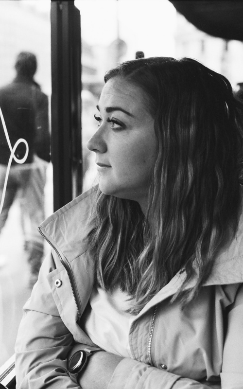 Black and white portrait of young woman gazing thoughtfully through window in jacket.