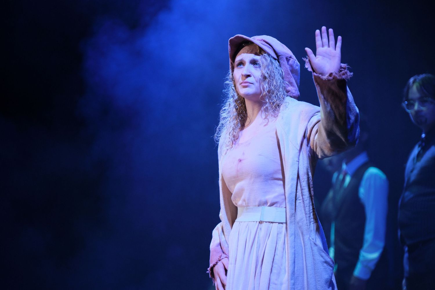 A theatrical performer in a white period costume raises her hand on a blue smoke-filled stage.