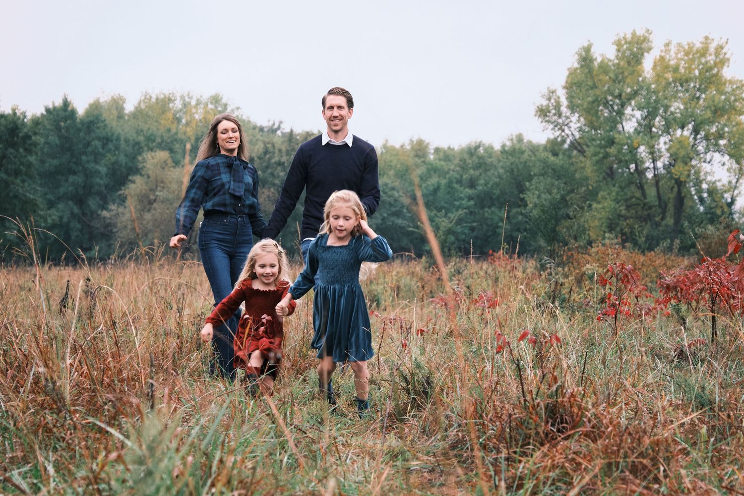 Family of four walking through autumn field with two young girls holding hands in front of parents.
