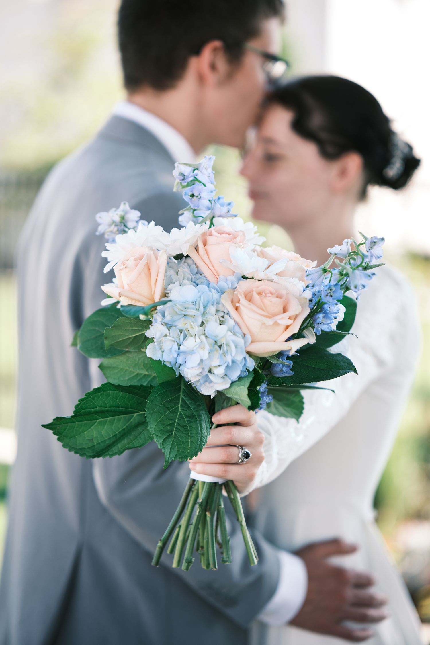 Close-up of a bridal bouquet with peach roses and blue hydrangeas held by a bride kissing her groom.