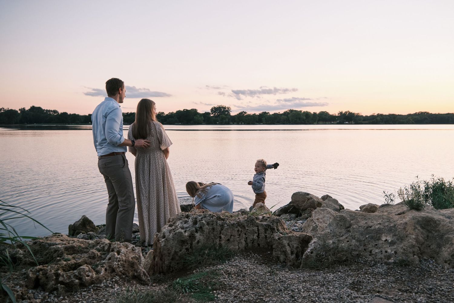 A family of four stands on rocky shores watching a serene lake at dusk during a maternity photo session.