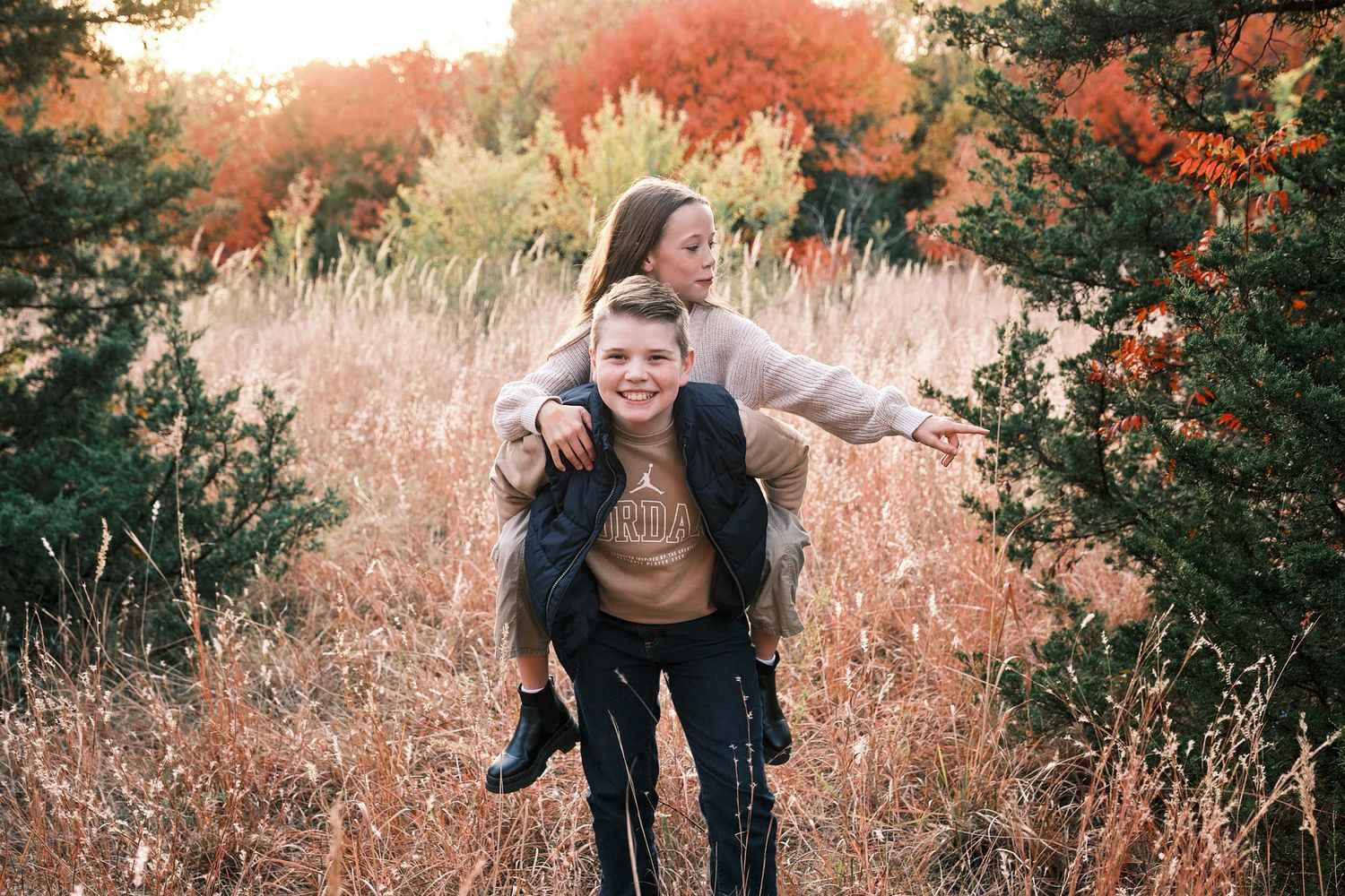 A smiling young man gives a laughing girl a piggyback ride through a golden autumn field surrounded by colorful foliage.
