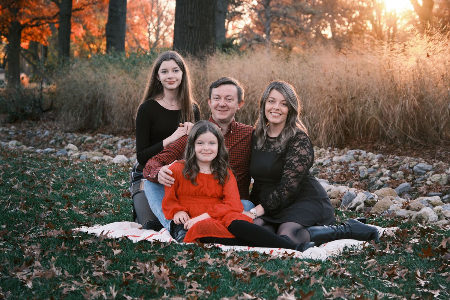 Happy family of four sitting on a blanket among fall leaves at golden hour in a park.