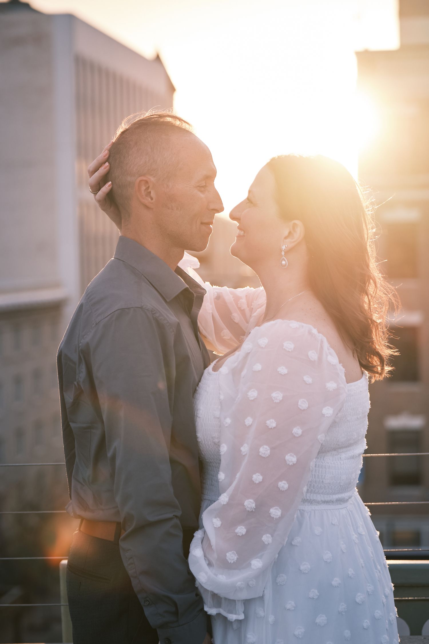 A couple shares a romantic moment on a rooftop at golden hour, the woman in a white polka dot dress.