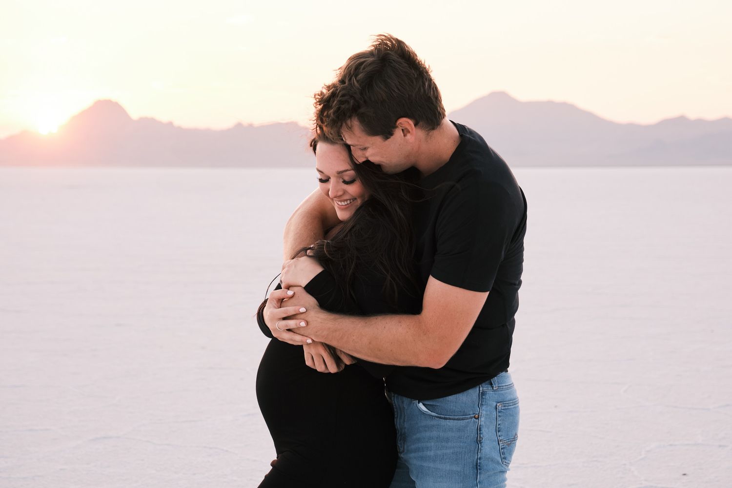Couple sharing tender maternity moment at sunset on white salt flats, man embracing pregnant woman.