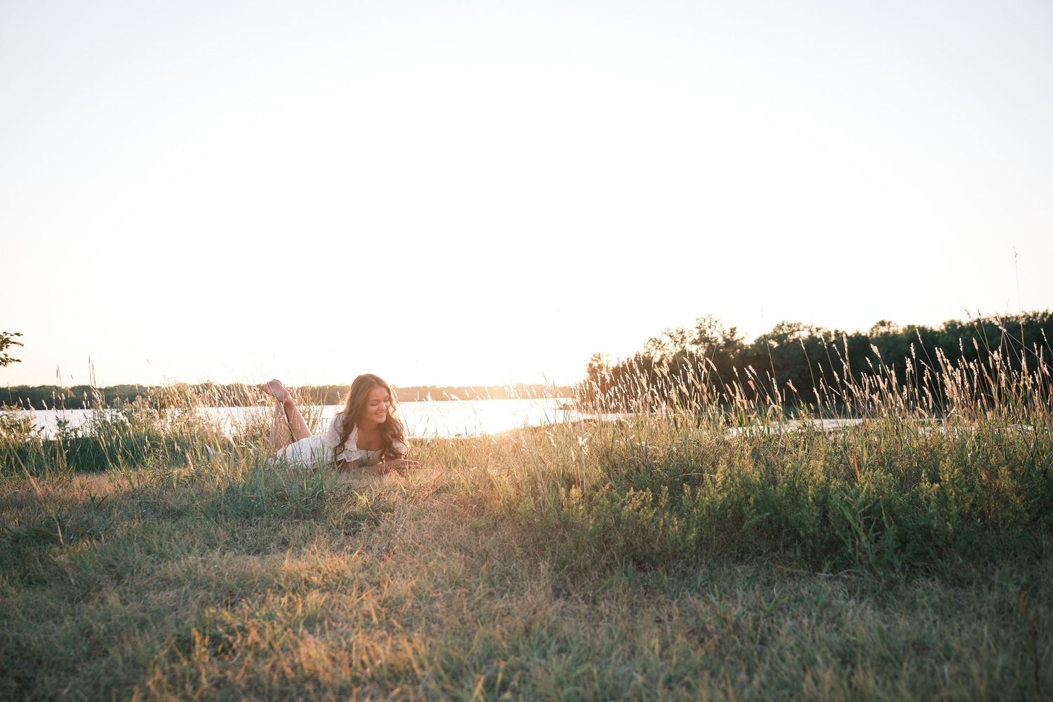 A young woman in a white dress sits peacefully in tall golden grass near a sunlit lake.