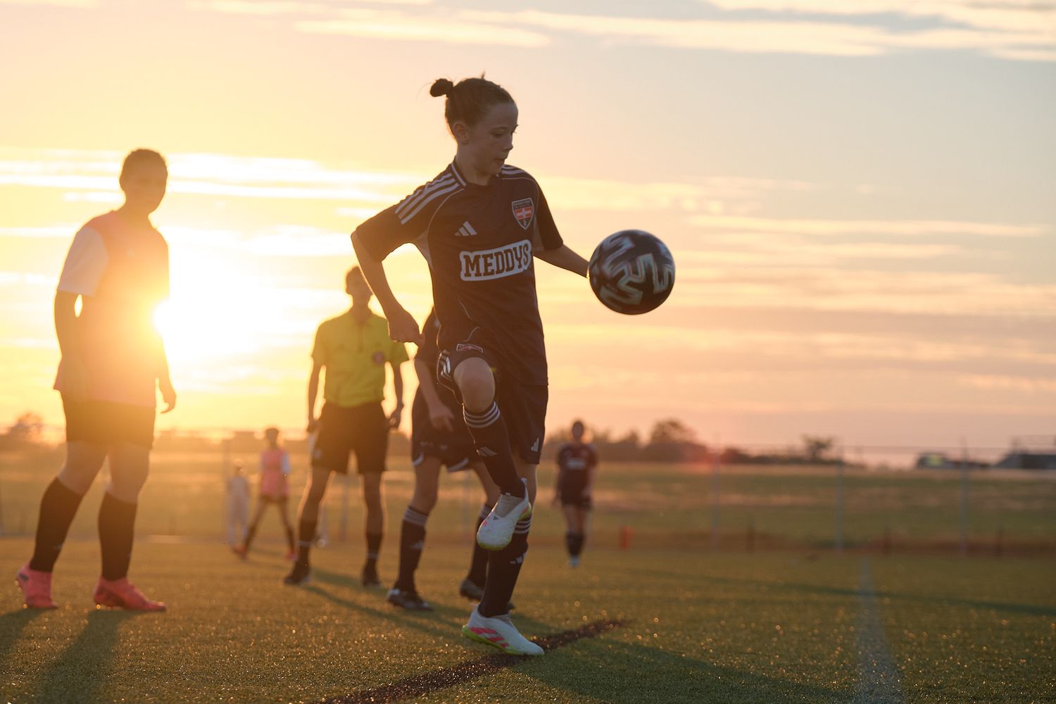 A soccer player controls the ball during a golden hour practice session on a green field.