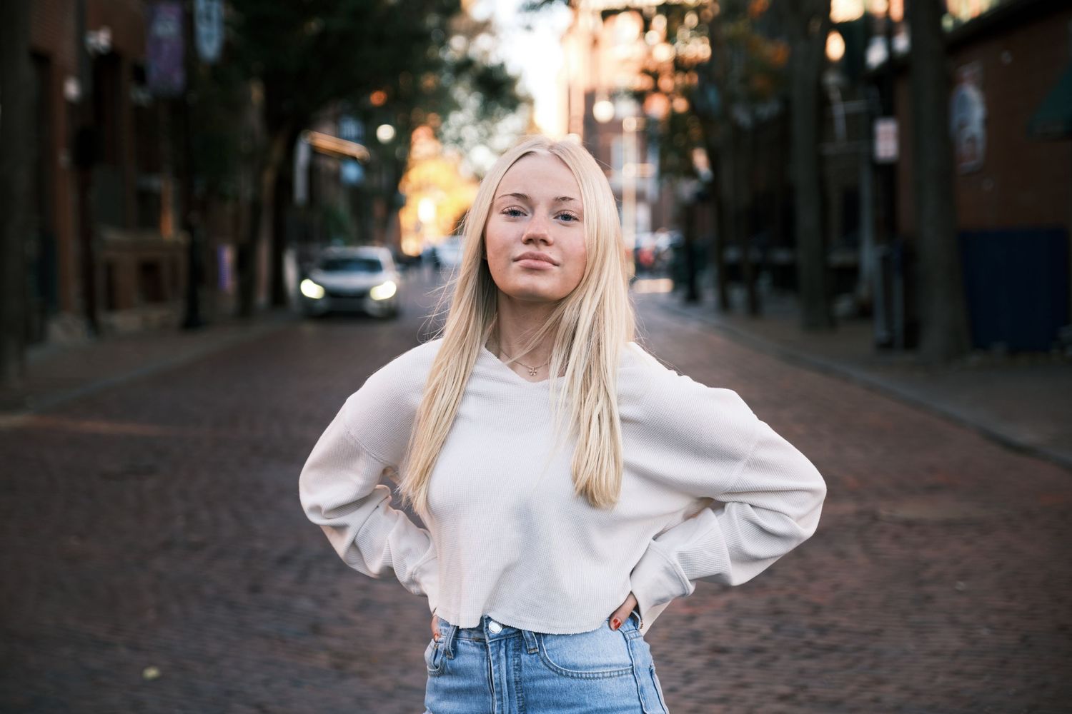 A confident blonde woman in a white sweater and jeans poses on a brick city street at dusk.