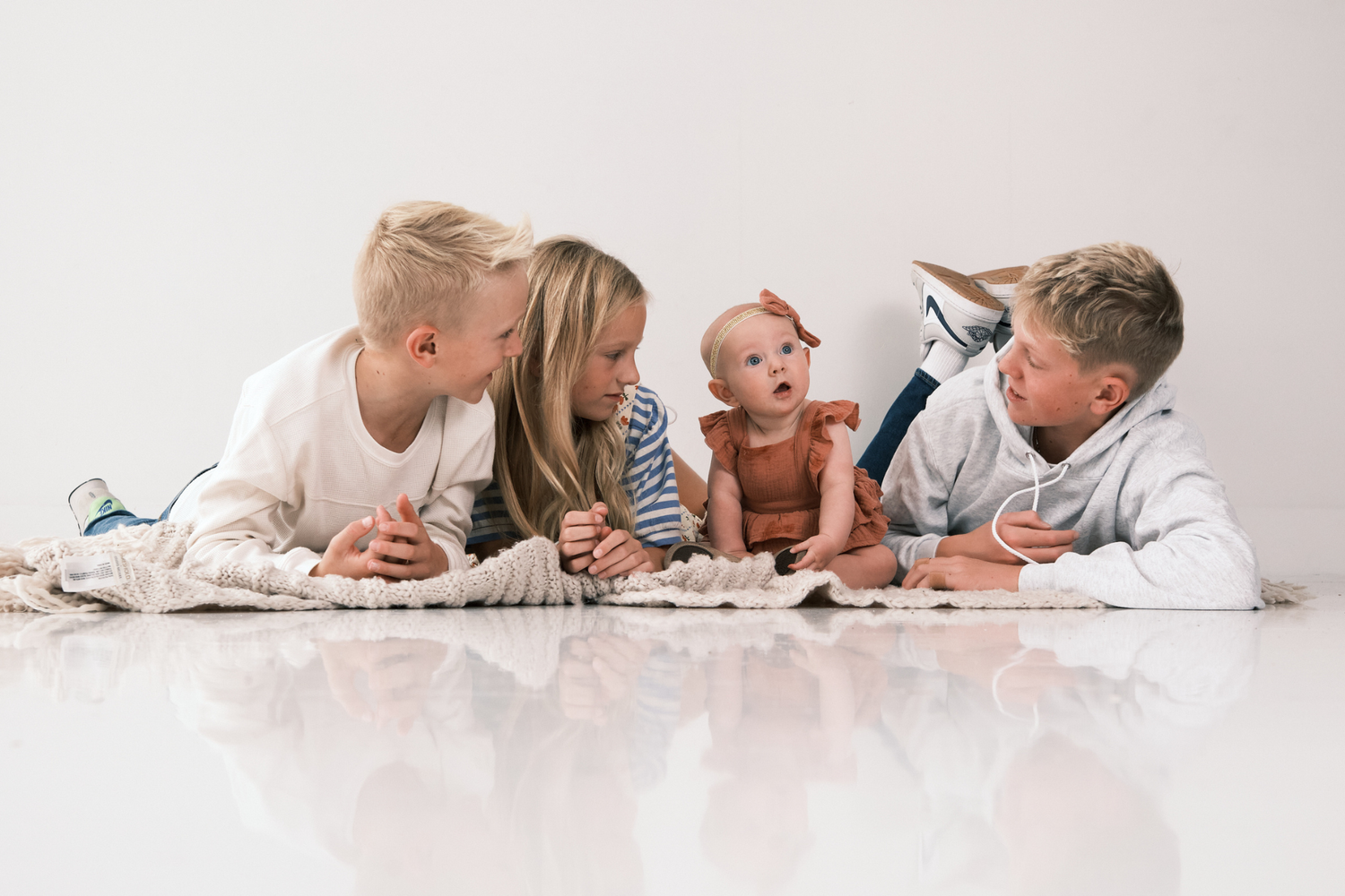 Three older siblings lie on the floor smiling at a curious baby during a bright white studio family photo session.