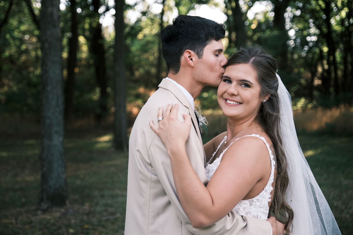 A bride in a white lace gown and veil smiles as her groom kisses her forehead in a wooded outdoor setting.