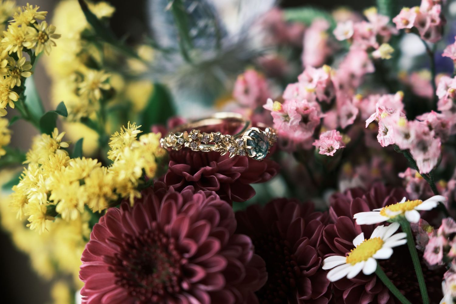 A delicate gold diamond engagement ring rests among colorful blooms including burgundy dahlias and yellow wildflowers.