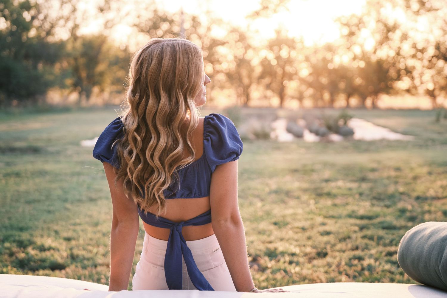 A young woman with long wavy blonde hair sits outdoors in a blue open-back top during a warm golden sunset.