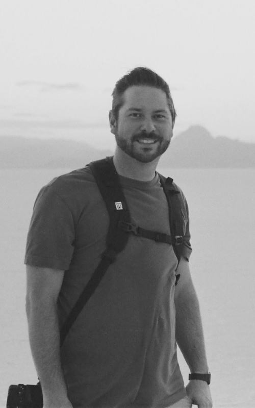 Head photographer Brian smiles with backpack and camera standing outdoors near a scenic landscape in black and white photo.