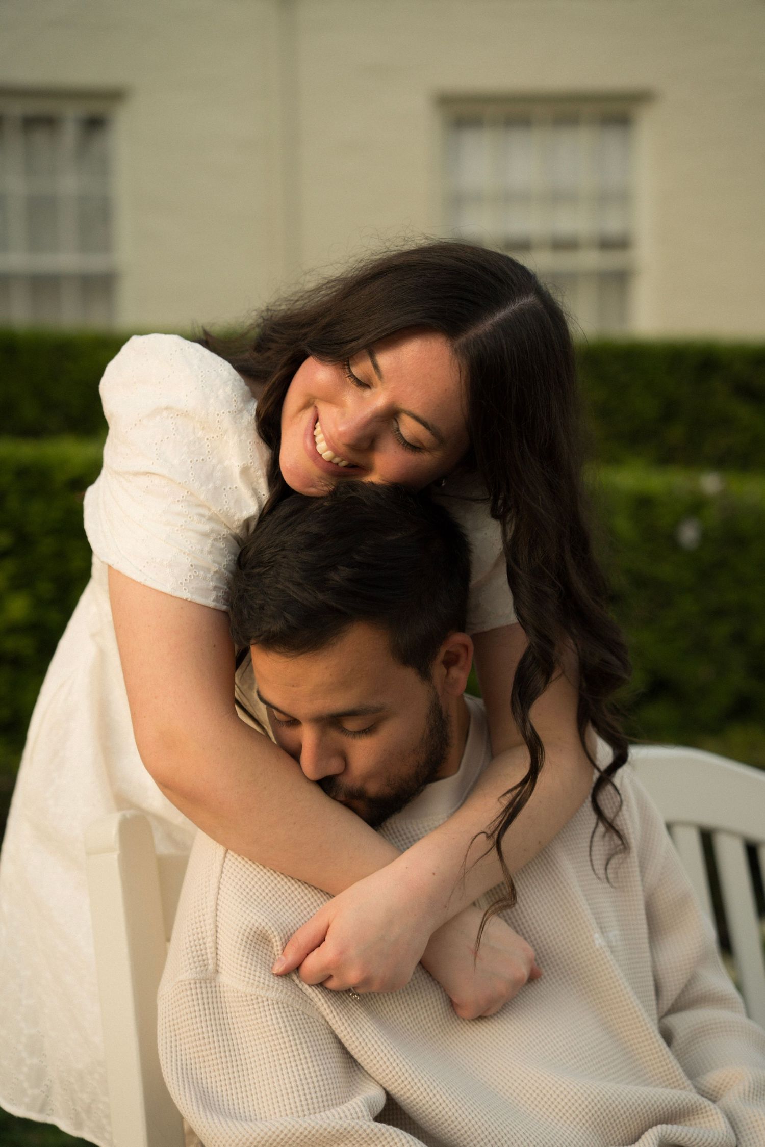 Wife standing over her husband leaning in with her head on his head and him kissing her arm
