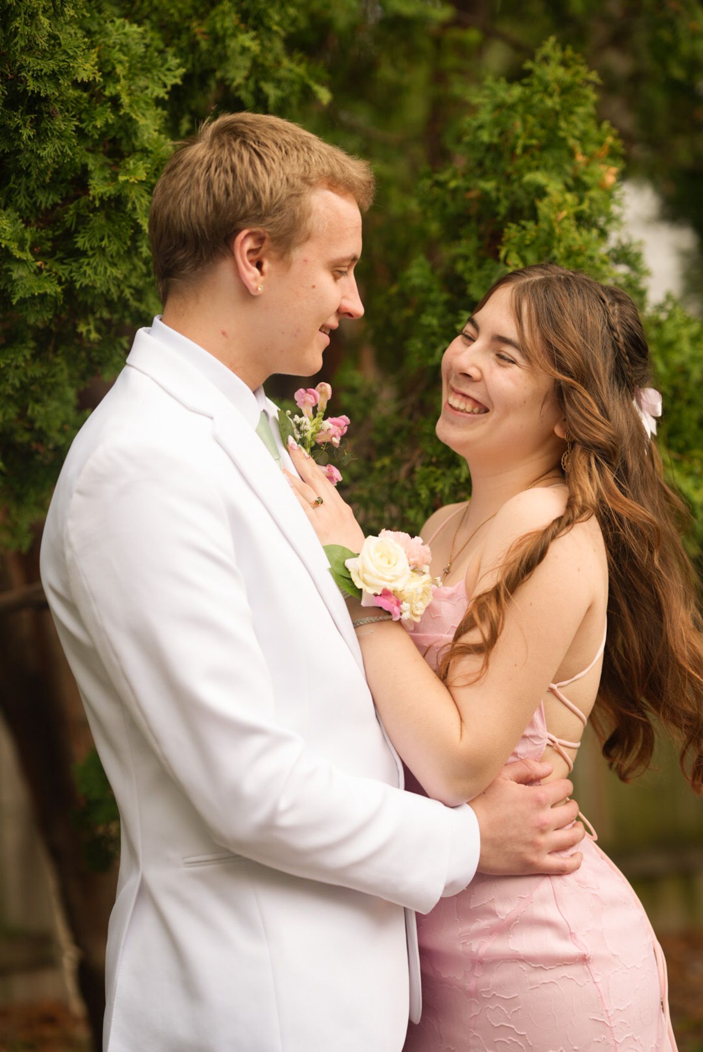 Smiling couple in pink dress and white suit with floral corsage posing together at prom outdoors.