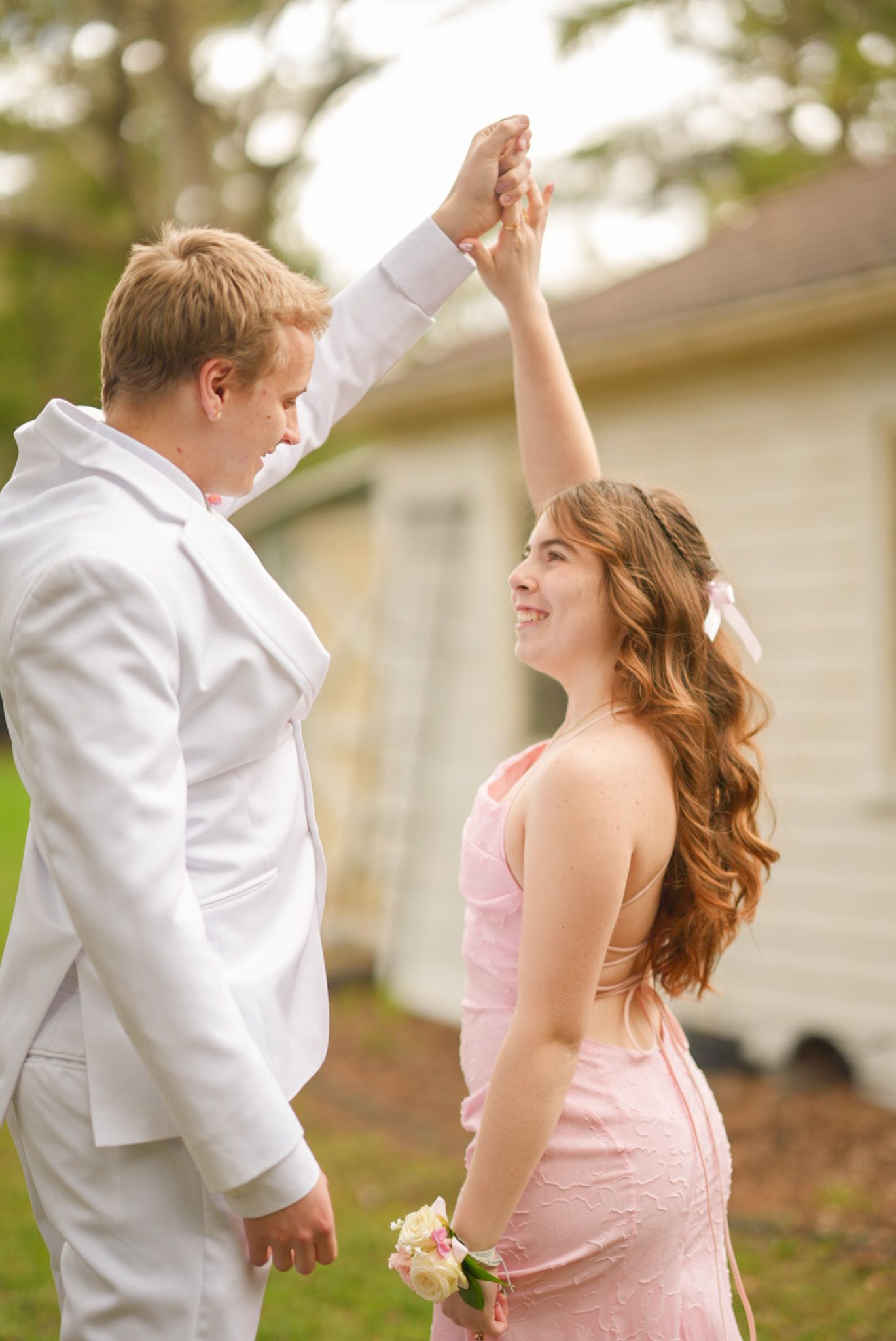 Smiling couple in pink dress and white suit with floral corsage posing together at prom outdoors.
