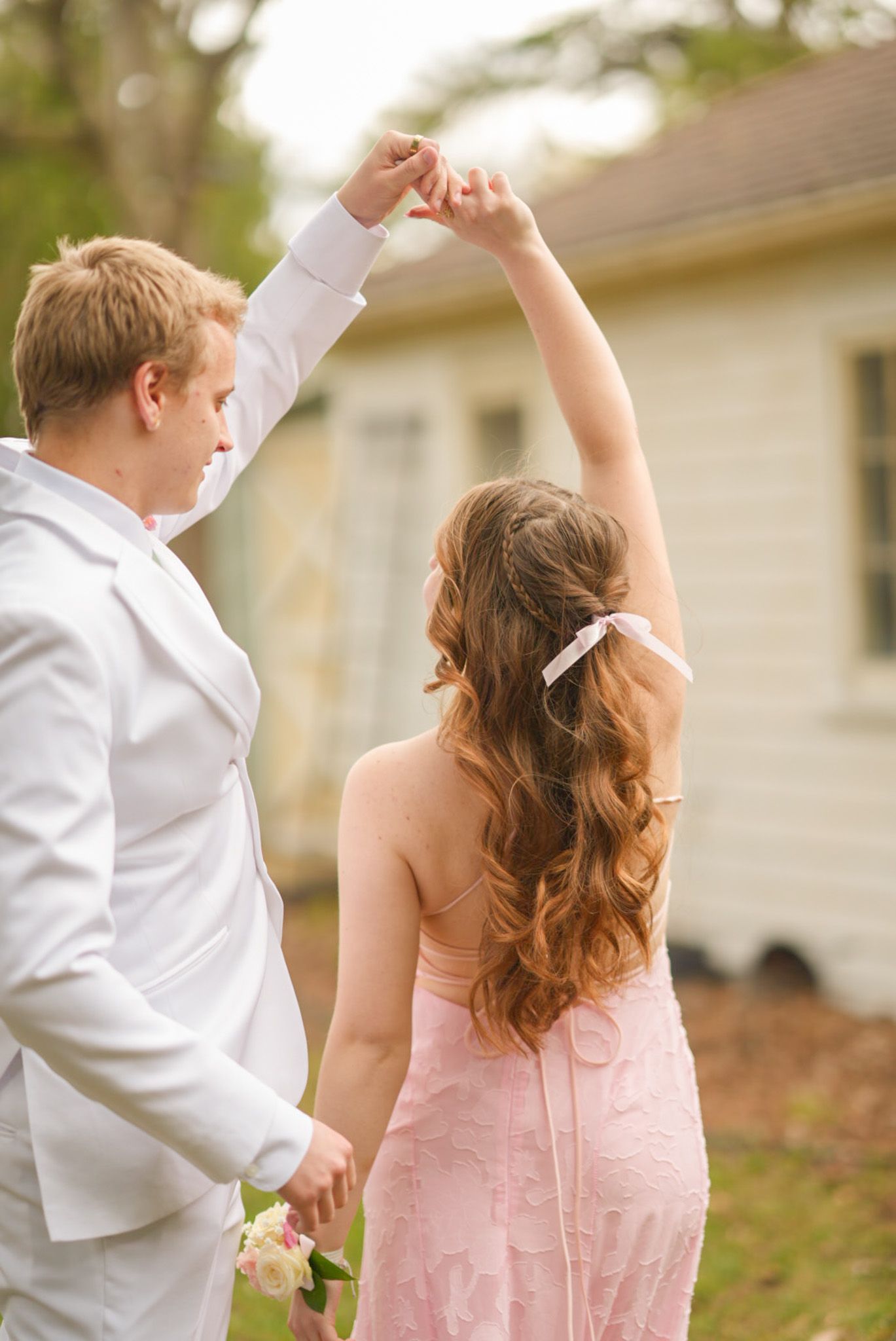 Smiling couple in pink dress and white suit with floral corsage posing together at prom outdoors.