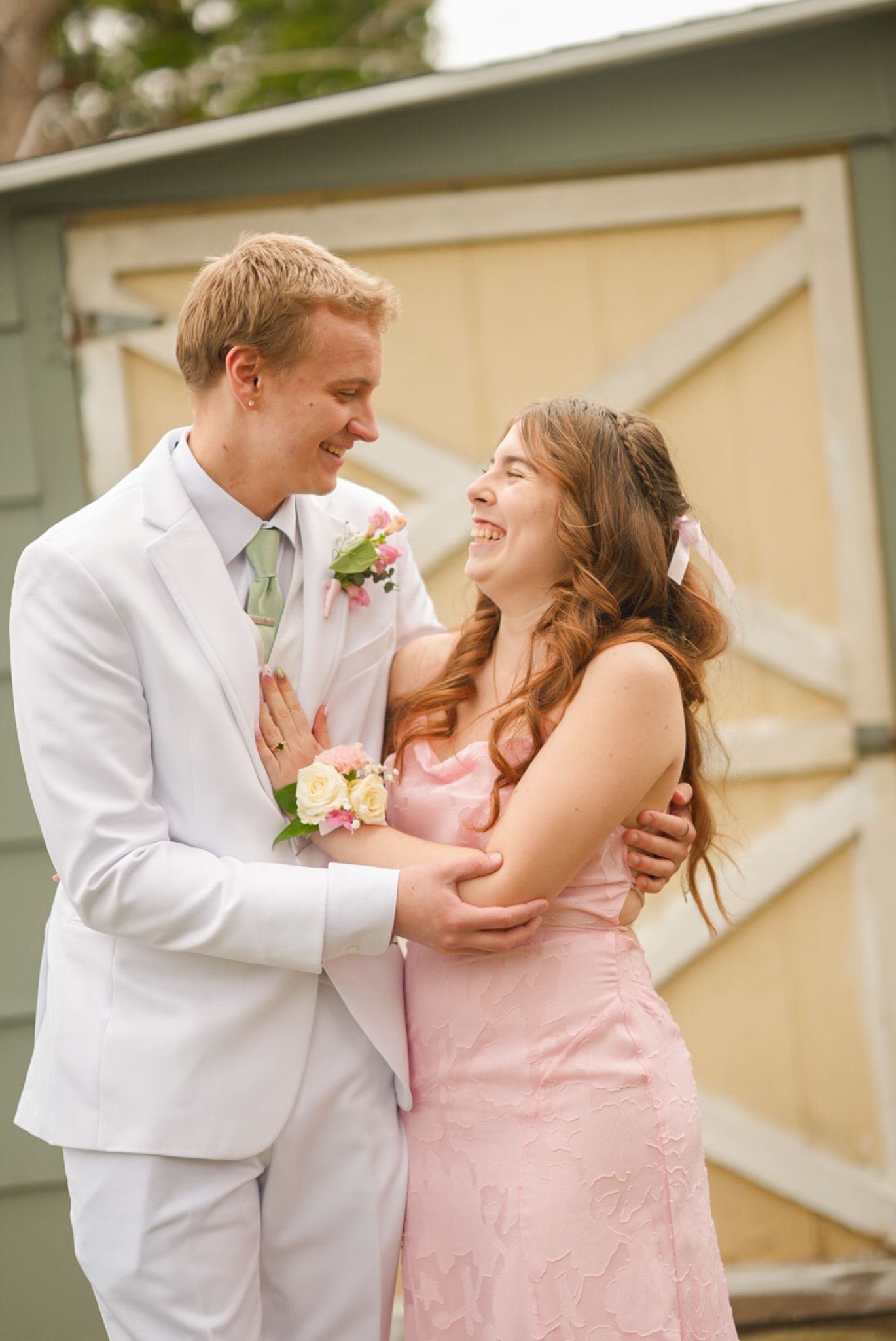 Smiling couple in pink dress and white suit with floral corsage posing together at prom outdoors.
