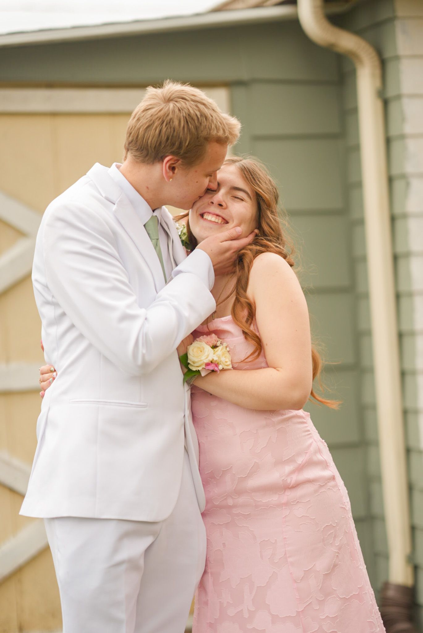 Smiling couple in pink dress and white suit with floral corsage posing together at prom outdoors.