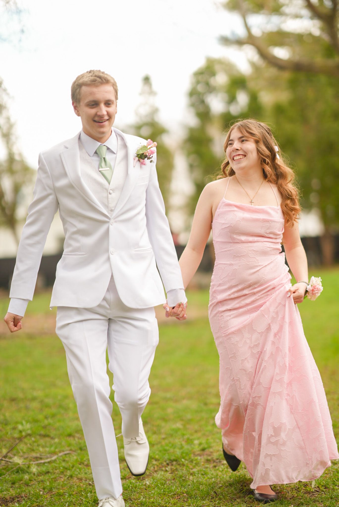 Smiling couple in pink dress and white suit with floral corsage posing together at prom outdoors.