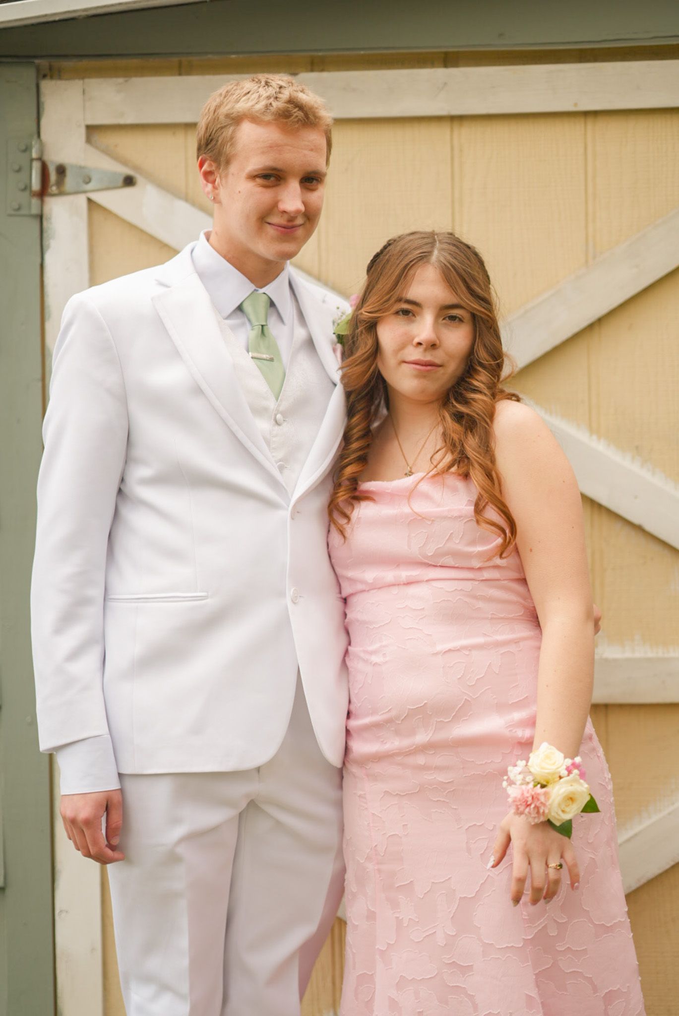 Smiling couple in pink dress and white suit with floral corsage posing together at prom outdoors.