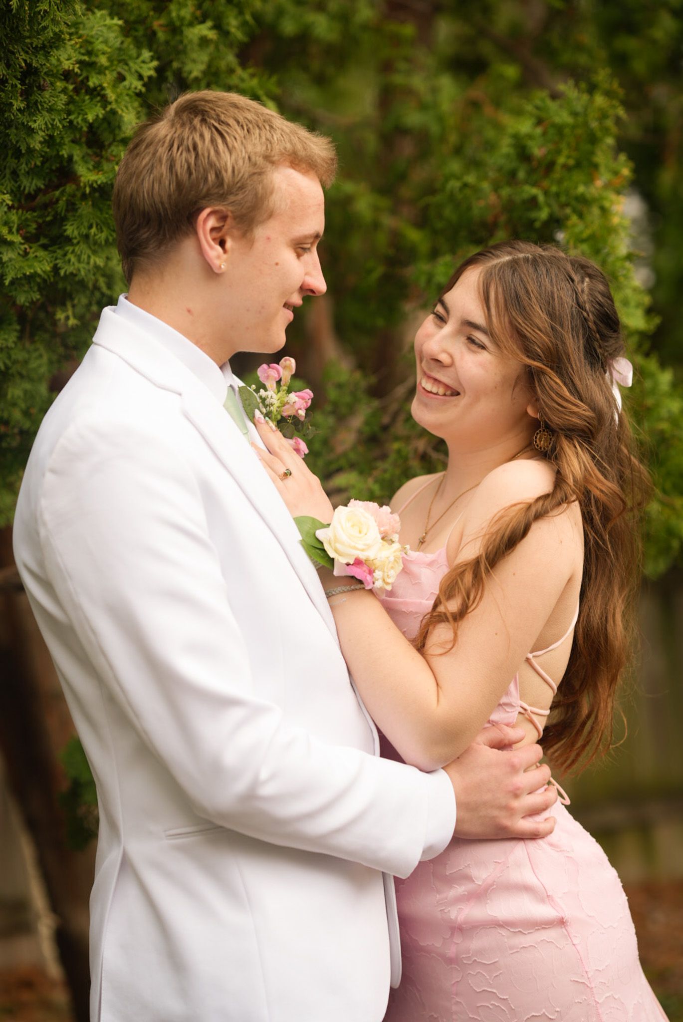 Smiling couple in pink dress and white suit with floral corsage posing together at prom outdoors.