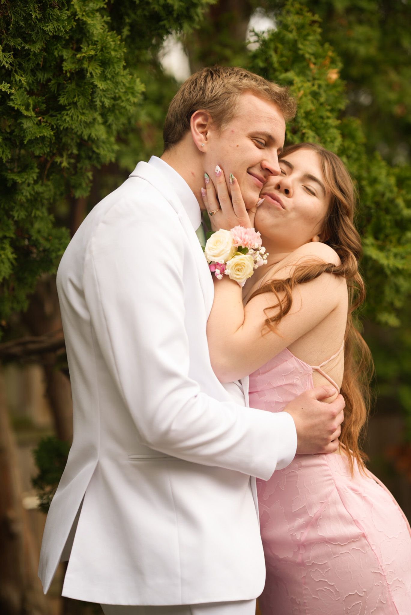 Smiling couple in pink dress and white suit with floral corsage posing together at prom outdoors.