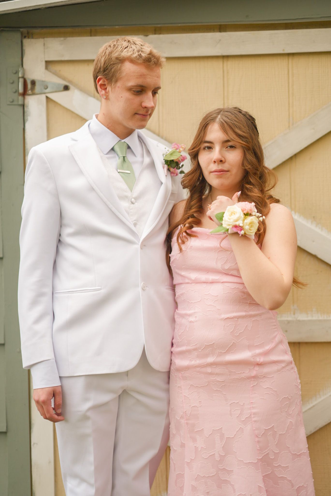 Smiling couple in pink dress and white suit with floral corsage posing together at prom outdoors.