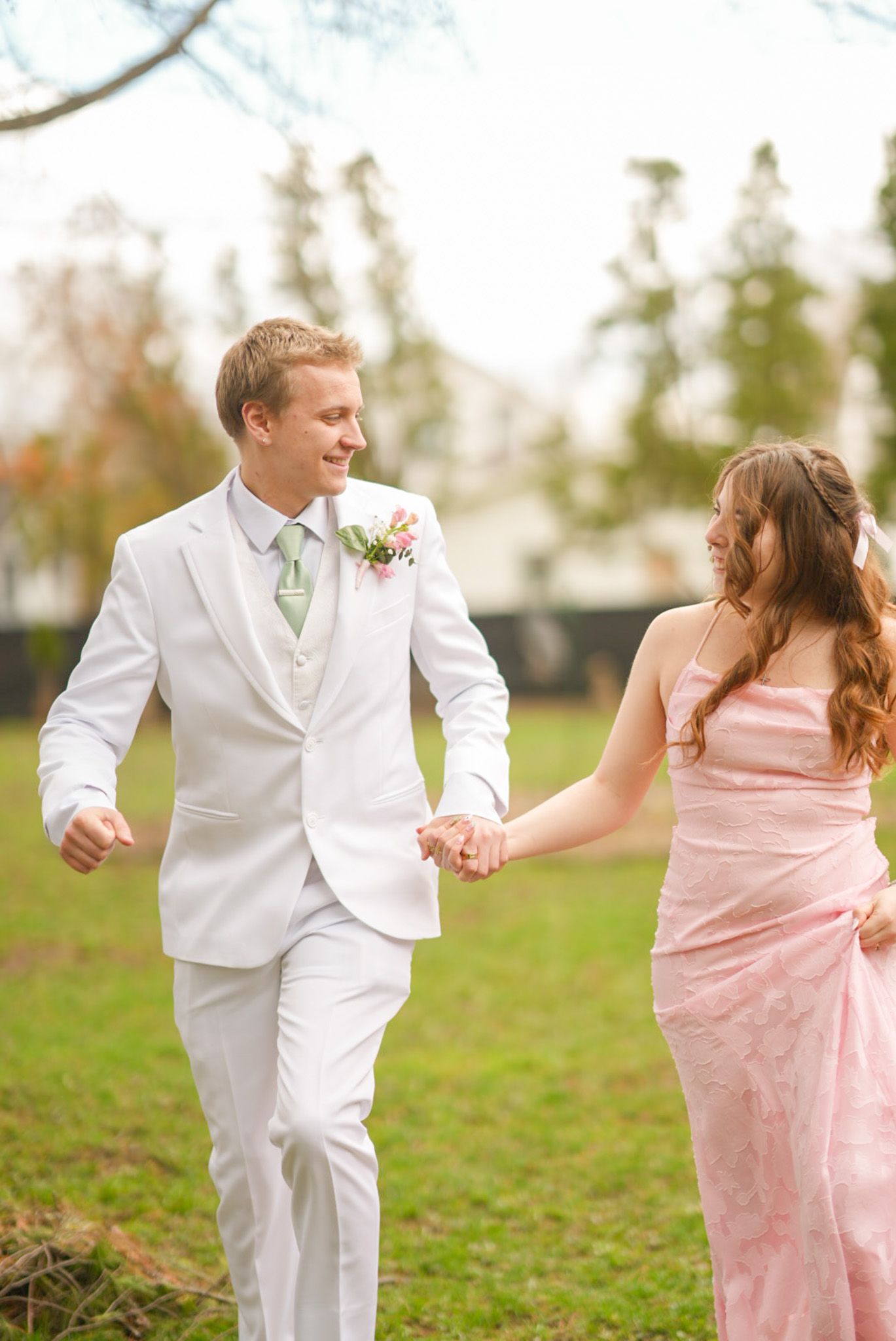 Smiling couple in pink dress and white suit with floral corsage posing together at prom outdoors.