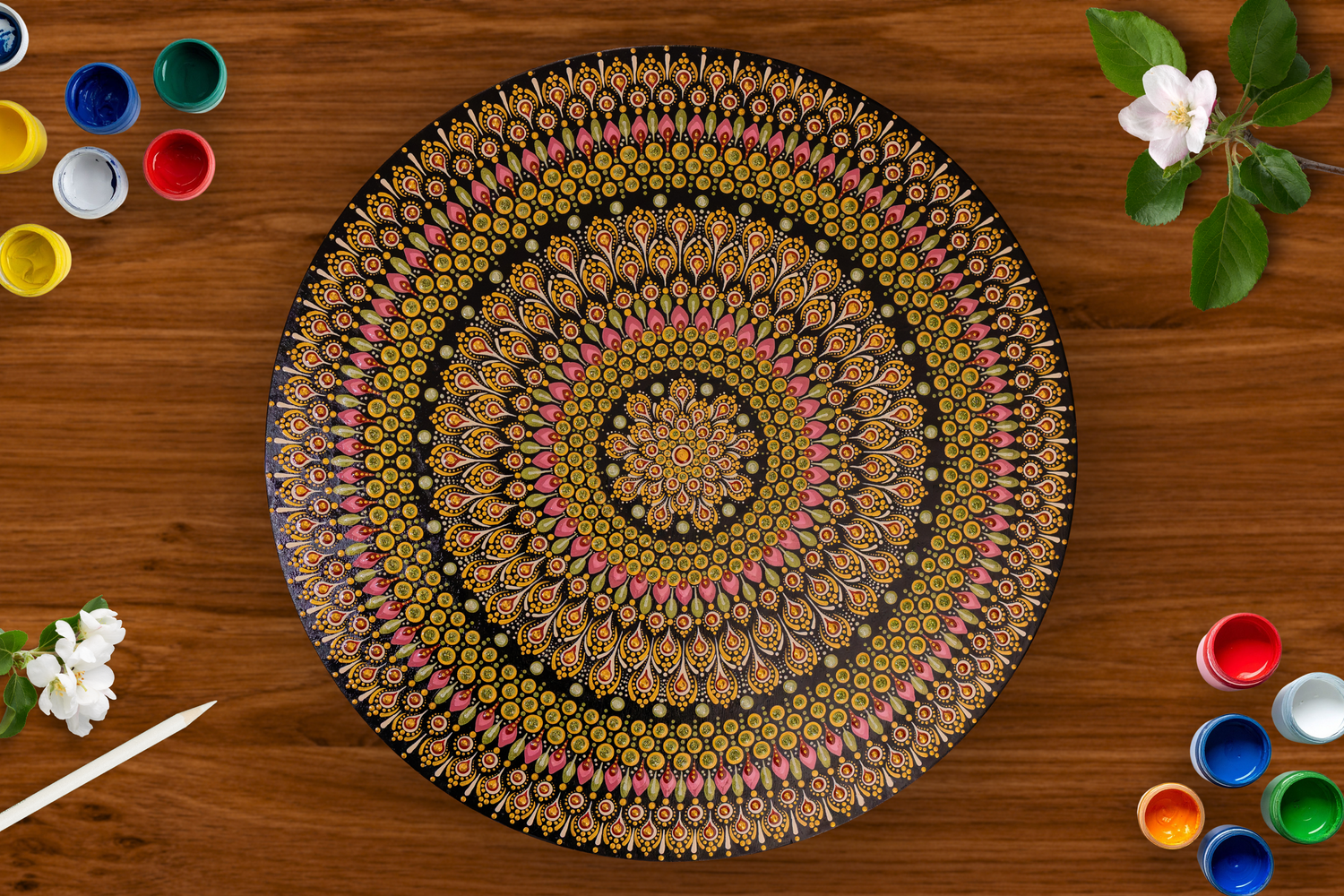Colorful hand-painted mandala dot art plate on wooden table with paint pots and white flowers.