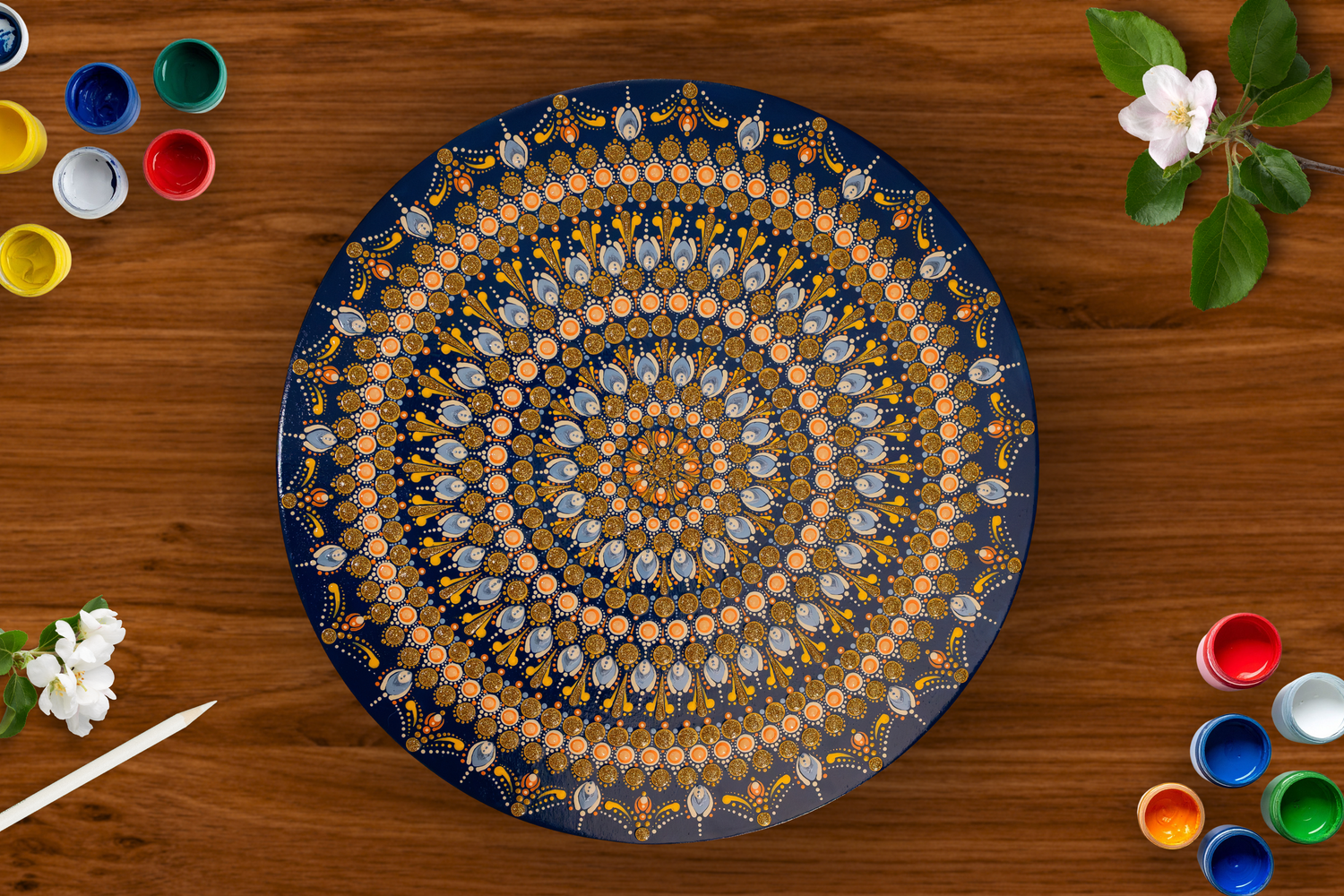 Handmade blue and gold mandala dot art decorative plate displayed on wooden table with paint pots and white flowers.
