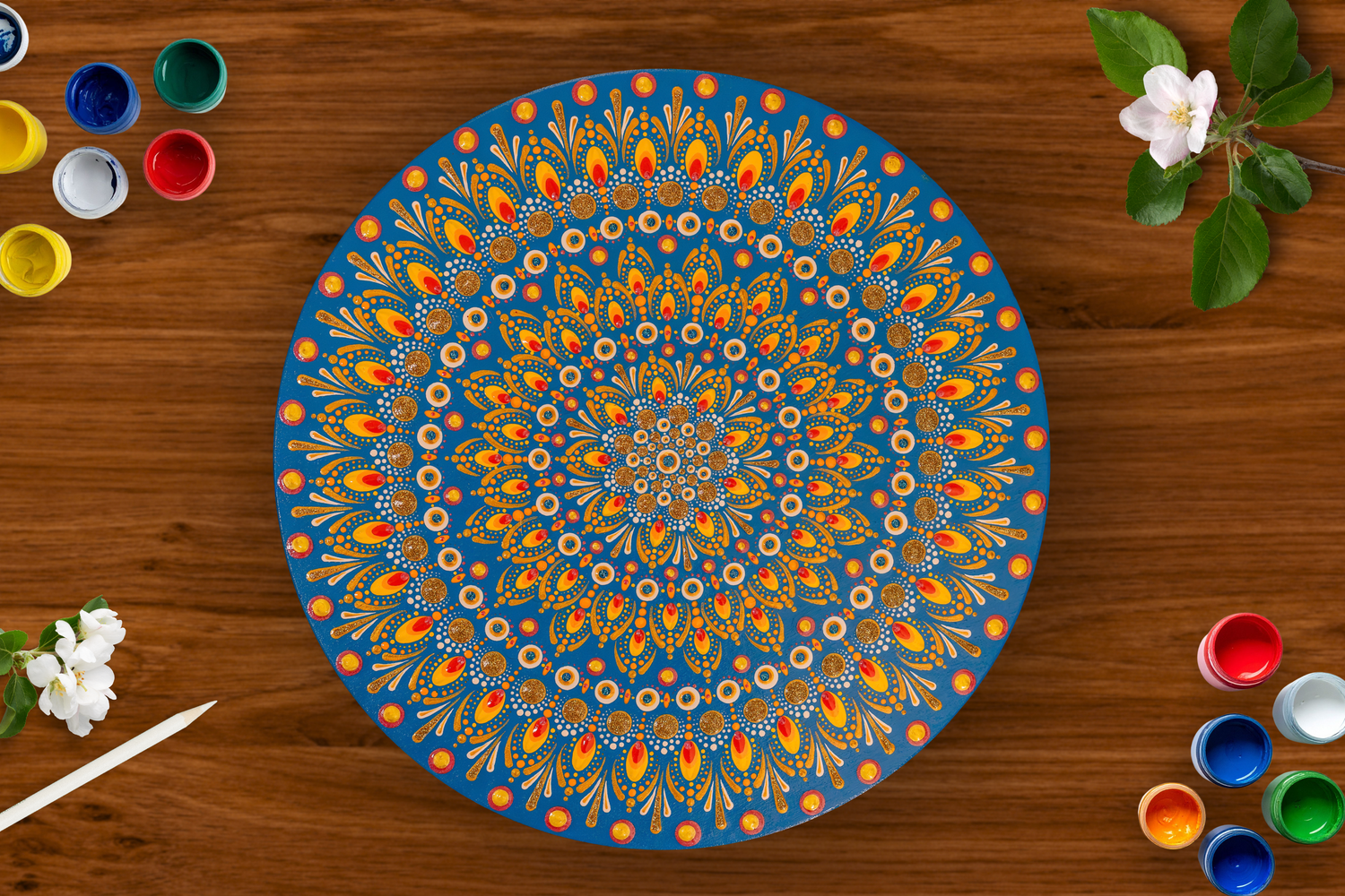 Colorful mandala dot painting on a blue plate surrounded by paint pots and white flowers on a wooden surface.