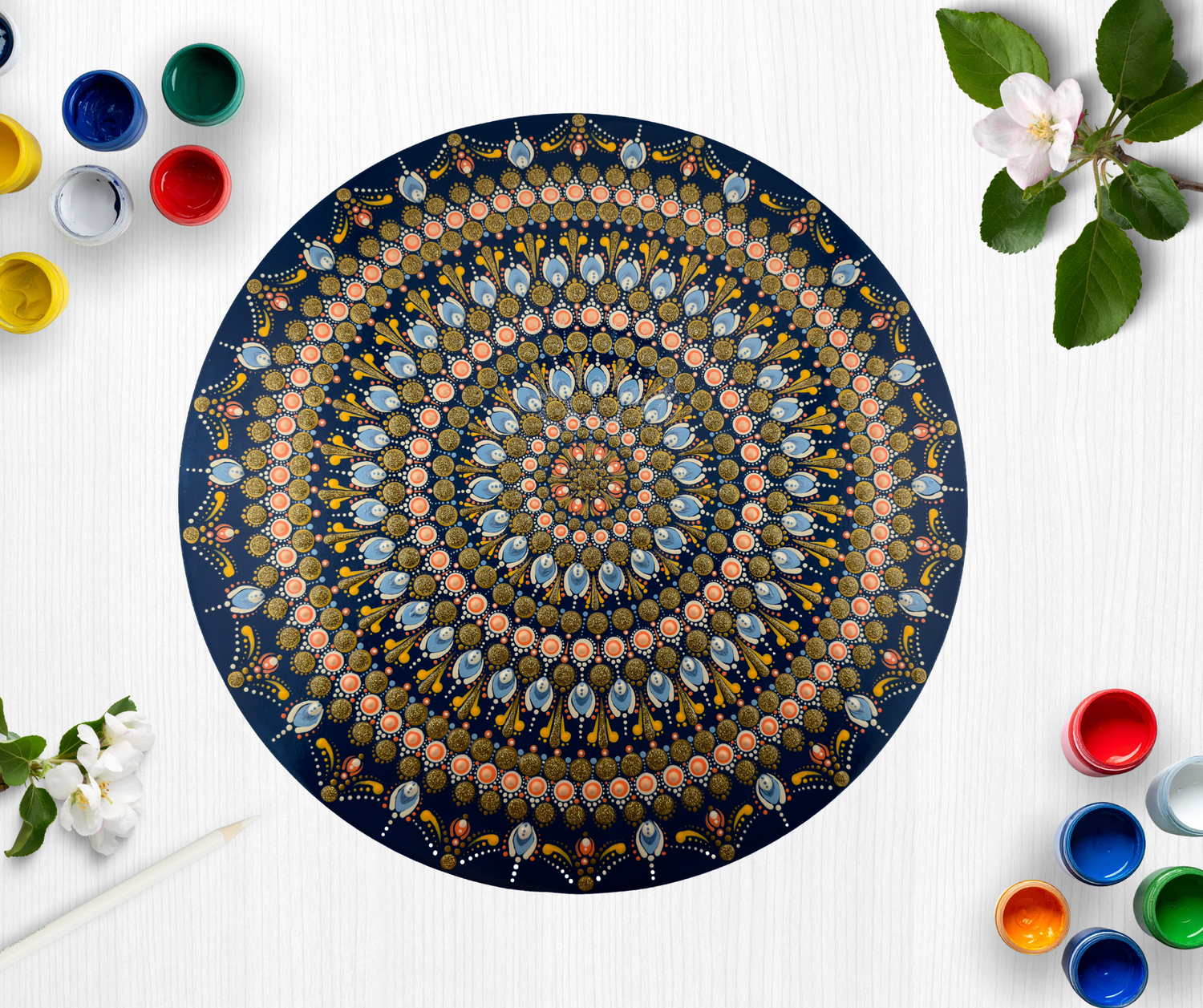 Intricate blue and gold mandala dot painting on round canvas surrounded by colorful paint pots and spring flowers.