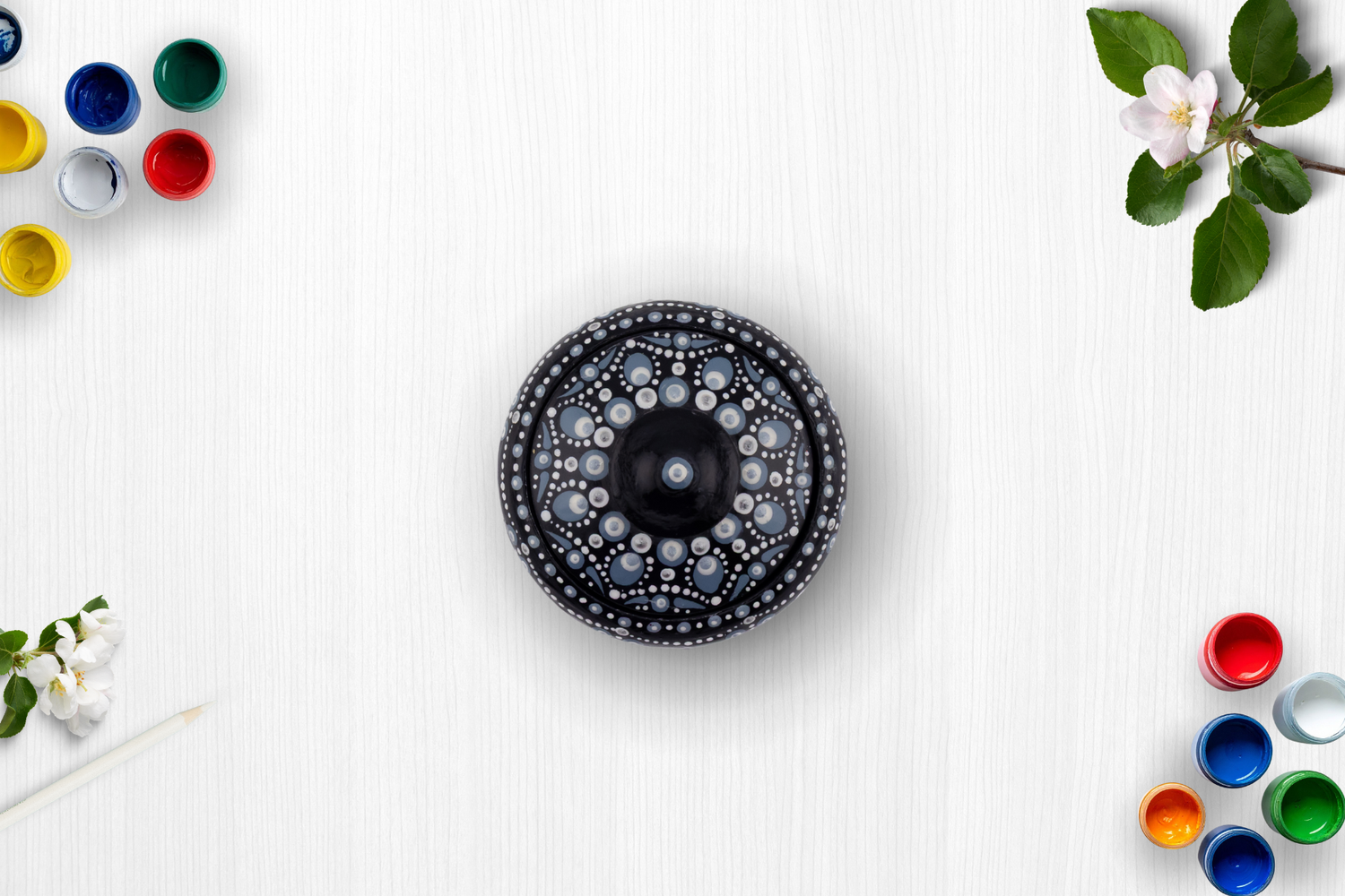 Black decorative yoyo with intricate white dot mandala pattern, surrounded by colorful paint pots and white flowers on white surface.