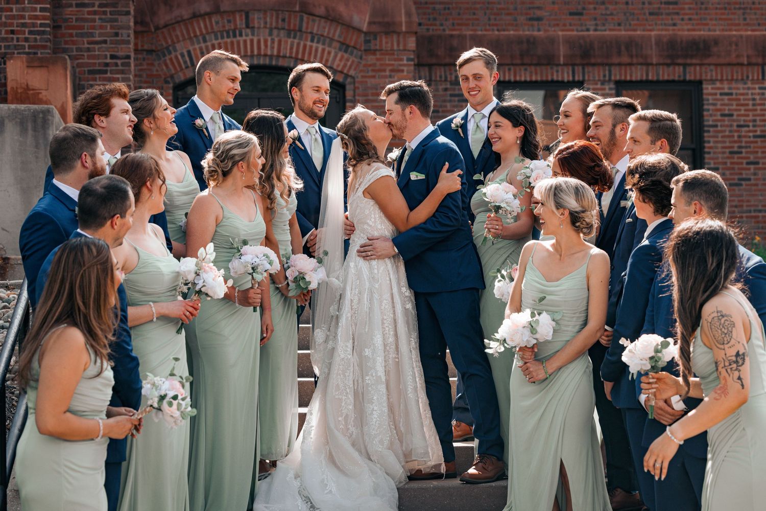 Wedding party in sage green gowns and navy suits gathered around couple sharing a kiss on steps.
