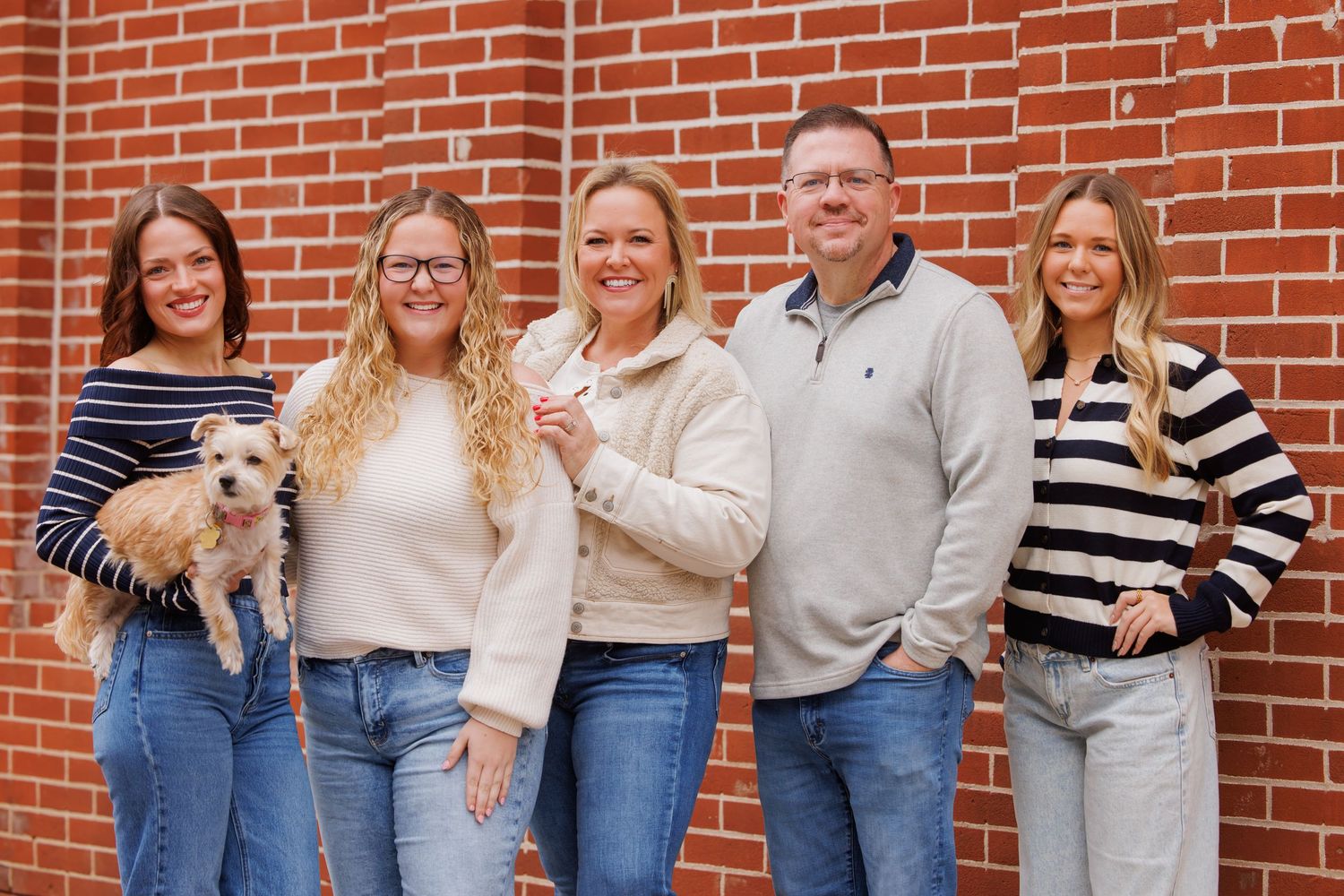 Smiling family of five with small dog posing together against a red brick wall backdrop.