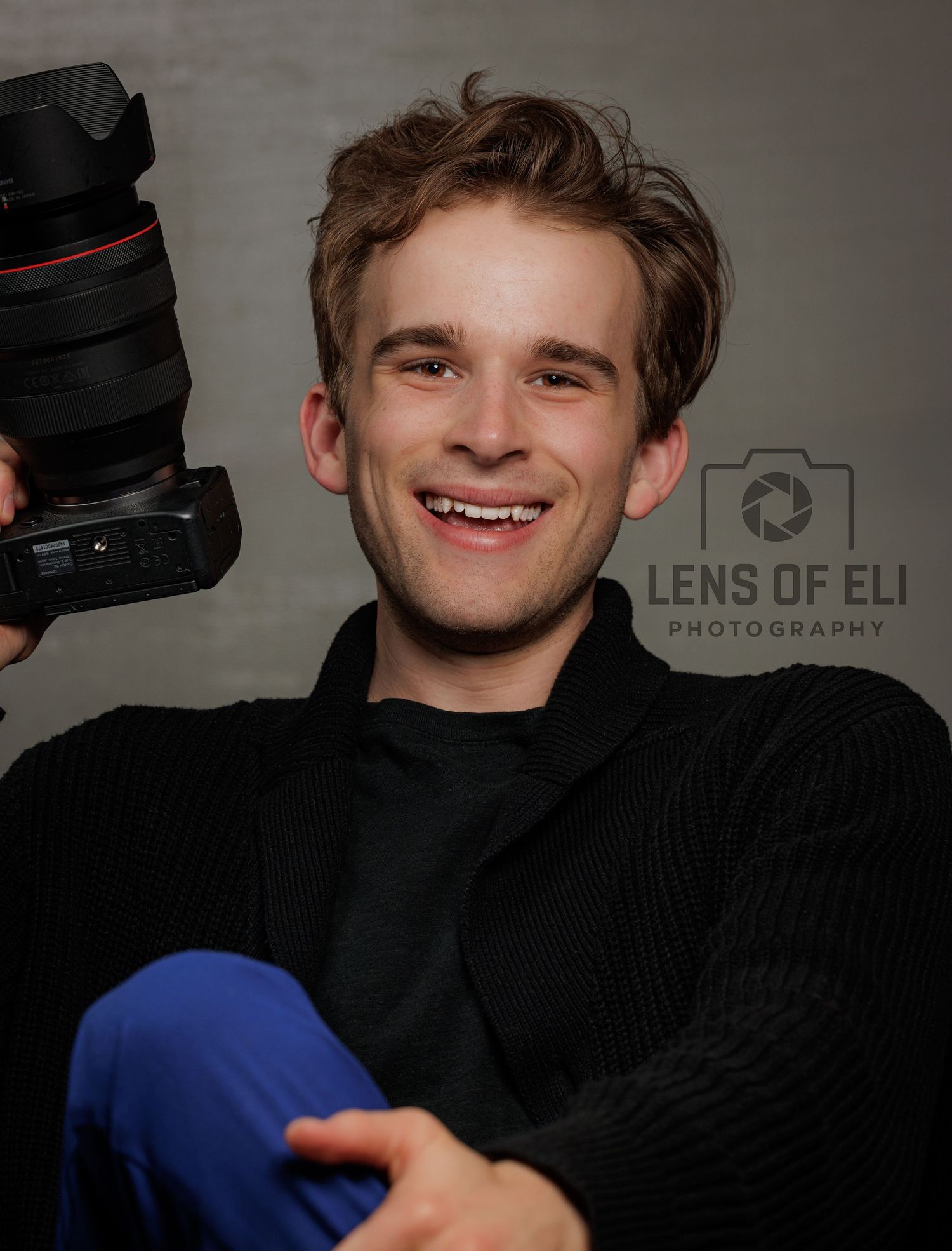 Young man smiling and holding a Canon camera, wearing a black jacket, promoting Lens of Eli Photography.