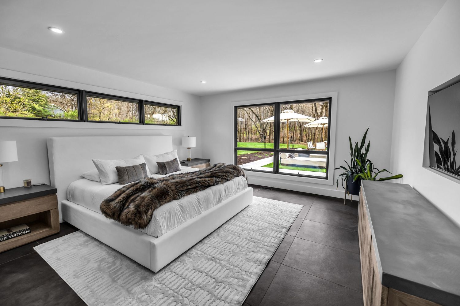 Modern master bedroom with white upholstered bed, dark tile floors, and floor-to-ceiling backyard view window.