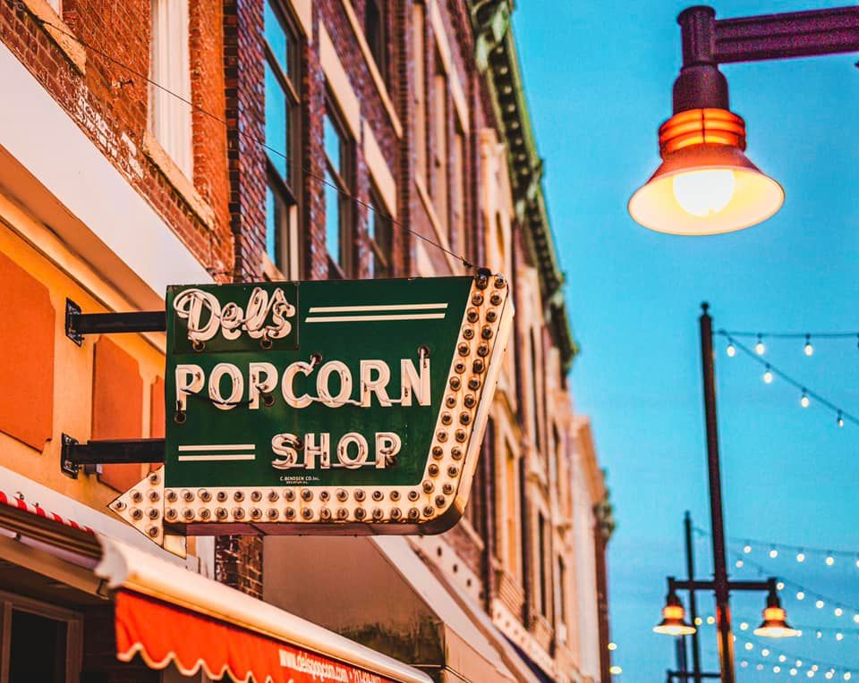 Vintage green Del's Popcorn Shop neon sign glowing at dusk on a charming historic brick street.