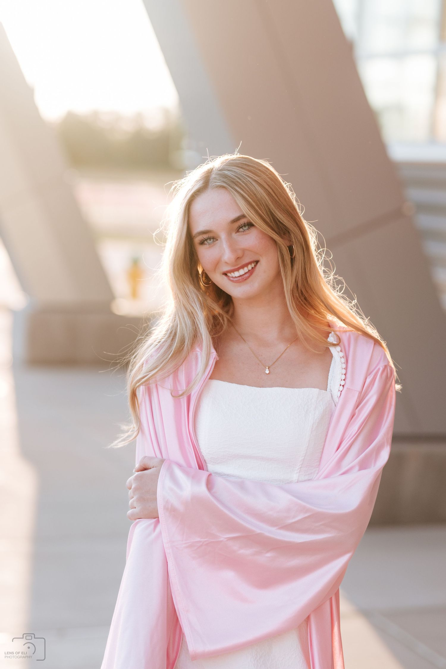 Smiling blonde woman in pink graduation robe posing outdoors in warm sunlight near modern architecture.