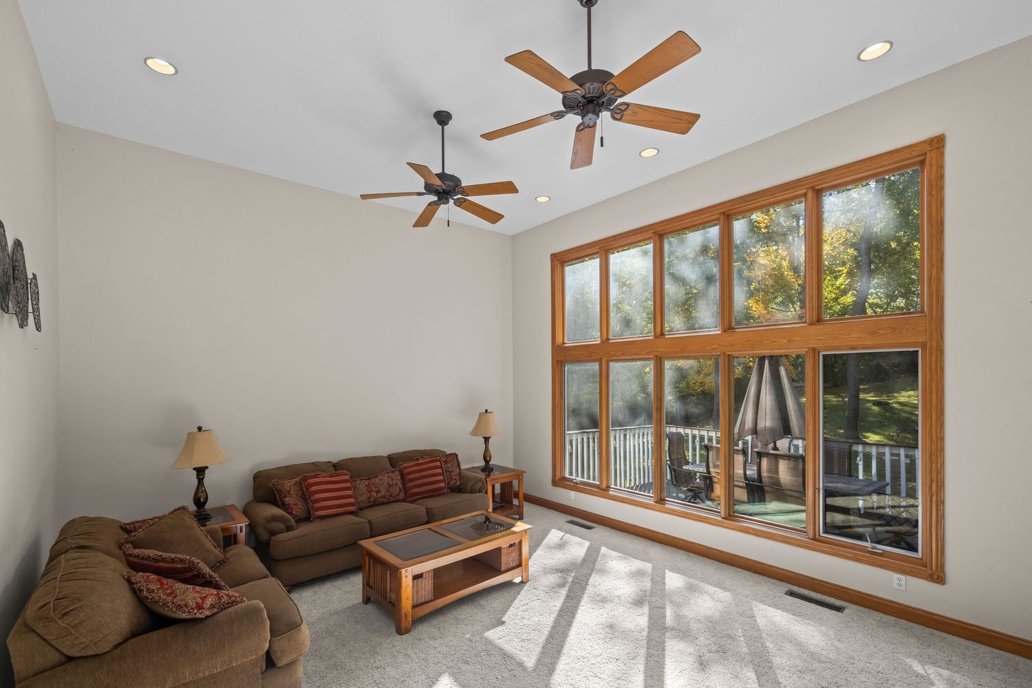 Bright living room with large wood-framed window, brown sofas, ceiling fan, and recessed lighting on light gray carpet.
