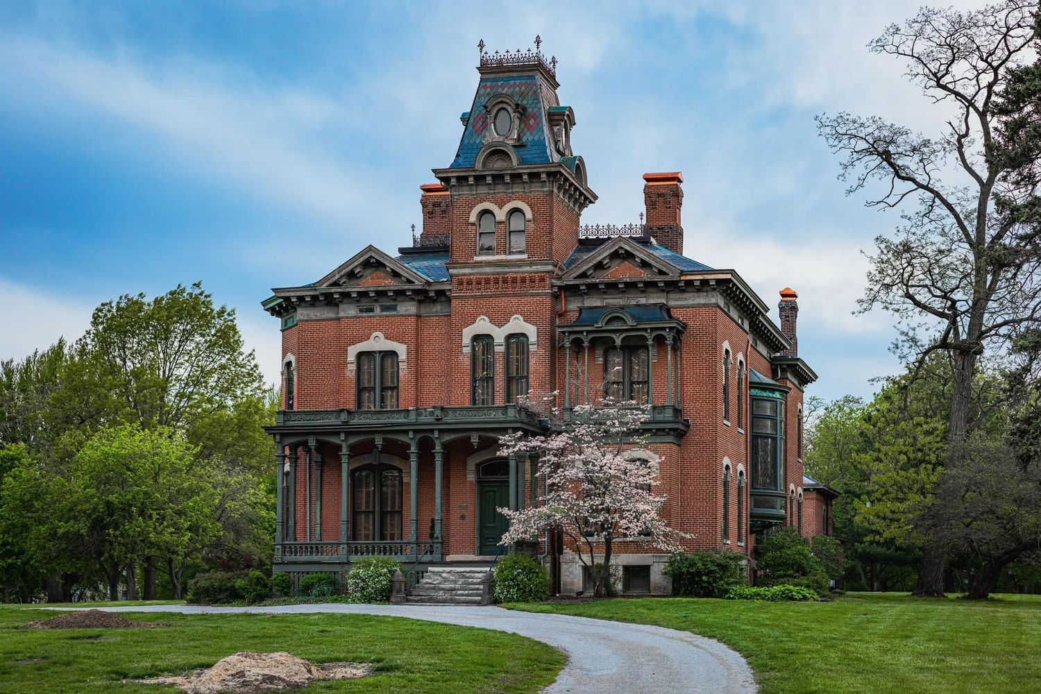 Victorian brick mansion with ornate details, tower, and blooming trees along a curved driveway.