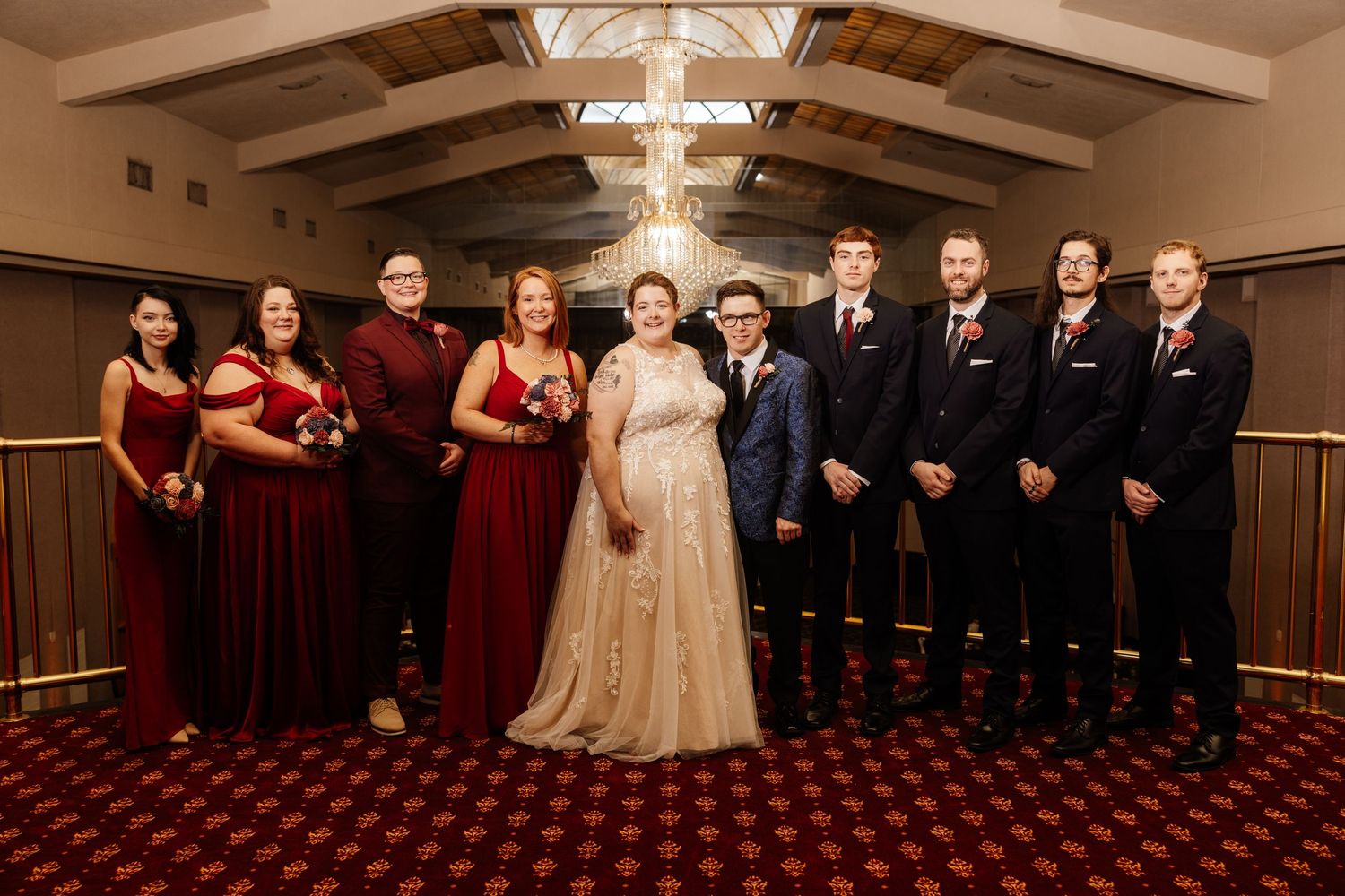 Wedding party posing in elegant venue, bridesmaids in burgundy gowns and groomsmen in navy suits flanking bride and groom.