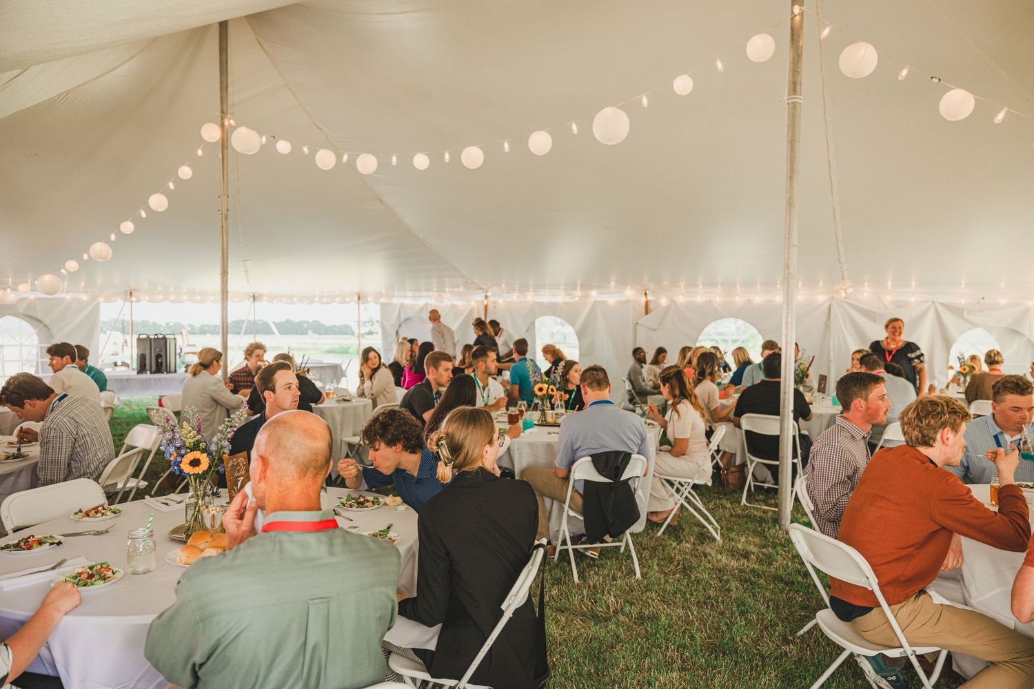 Guests dining under a white tent with string lights and paper lanterns at an outdoor reception.