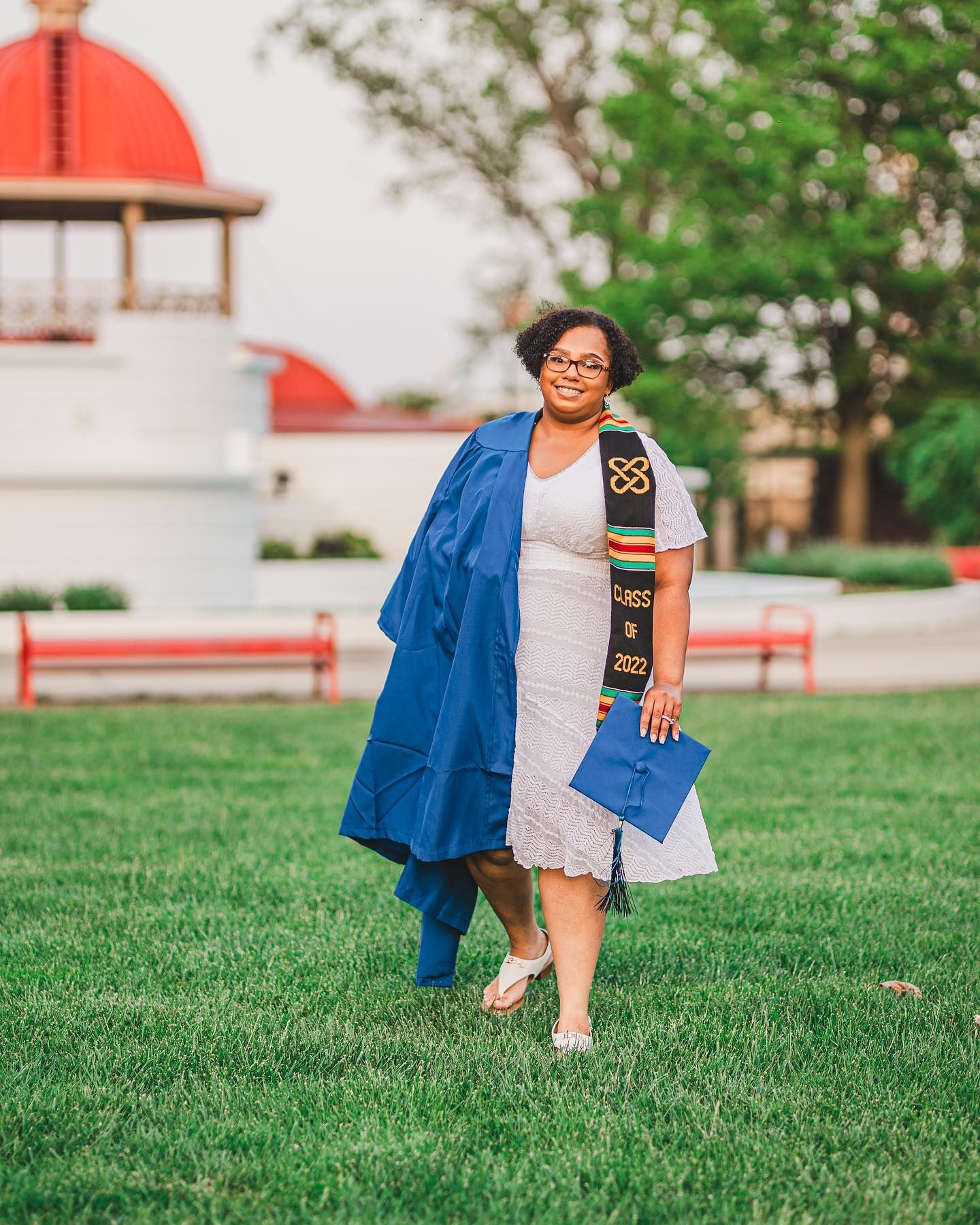 Woman in blue graduation gown and cap holding diploma on green campus lawn with red-roofed building in background.